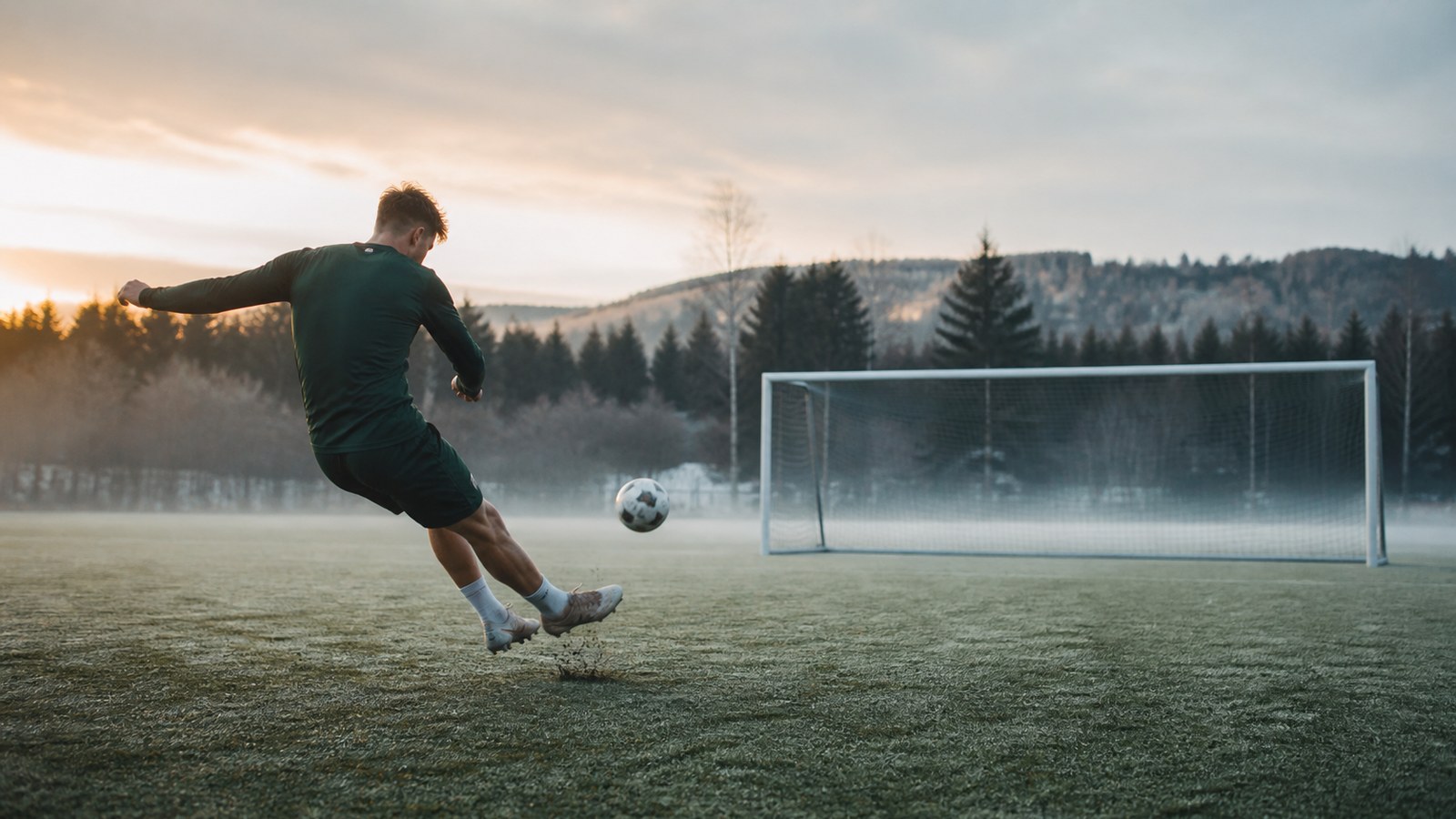 Powerful soccer striker in plain dark green kit shooting toward an empty goal on a cold morning